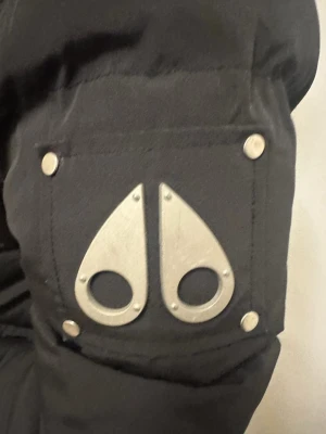 Svart dunjacka med päls från Moose Knuckles - Svart dunjacka från Moose Knuckles med ikonisk metall-logga på ärmen, fluffig avtagbar huva med ljus päls, dragkedja och knappar framtill samt flera fickor. Jackan har en schysst puffer-look och är perfekt för kalla dagar.