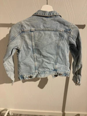 Ljusblå jeansjacka med knappar - Snygg ljusblå jeansjacka med klassisk krage och silverfärgade knappar framtill. Jackan har två bröstfickor med lock och knapp, samt justerbara knappar vid midjan. Perfekt för dig som gillar en chill och tidlös stil.