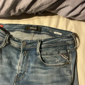 Replay Anbass ljusblå jeans, stl 30/32 - Säljer ett par Replay Anbass jeans i ljusblå tvätt med snygga slitningar och klassiska fem fickor. Jeansen har normal passform och raka ben, med Replay-logga på fickan och svart läderpatch bak. Perfekt för dig som gillar stilrena och bekväma jeans. Litet håll där på benet men inget som syns och som man lätt kan dra igen med nål och tråd