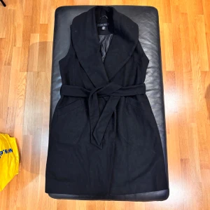 Svart omlottväst med bälte från Amisu - Svart lång väst från Amisu med bred krage och omlott-design. Västen har ett matchande bälte i midjan och två fickor framtill. Materialet är mjukt och känns som ullblandning, perfekt för lager-på-lager. Snygg och stilren look som lyfter din outfit.