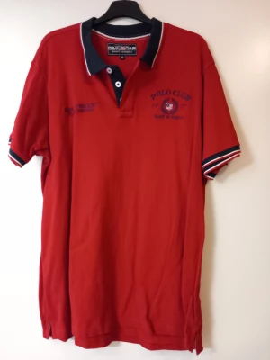 Röd pikétröja Polo Club Saint Vincent XL - Röd pikétröja från Polo Club Saint Vincent i storlek XL. Tröjan har broderade detaljer på bröstet, klassisk krage i svart med vit kant och korta ärmar med randiga muddar. Materialet är bomull och passformen är normal. Oanvänd.
