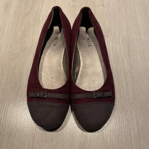 Vinröda ballerinaskor från Bonita - Snygga ballerinaskor i vinröd mocka från Bonita med mörkgrå tå och dekorativt band med liten spänne framtill. Skorna har platt sula och rund tå, vilket gör dem både stilrena och bekväma. Perfekta för dig som vill ha en klassisk men ändå trendig look.
