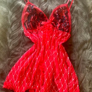 Röd topp med volang och rosett - Supersöt röd topp  i mesh med tunna axelband, volangdetaljer framtill och liten rosett vid bysten. Överdelen har broderade rosor i svart och kjolen har ett rutigt mönster i vitt. Perfekt för dig som vill sticka ut med en lekfull och romantisk look.