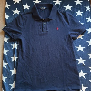 Marinblå pikétröja från Ralph Lauren - Klassisk marinblå pikétröja från Ralph Lauren med röd broderad logga på bröstet. Tröjan har korta ärmar, krage och två knappar framtill. Tillverkad i mjuk bomull som är skön mot huden. Perfekt för en stilren och avslappnad outfit. Är i storlek XL för barn!! Så passar en som har storlek S i herr 