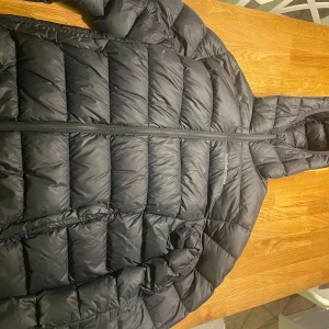 Peak Performance jacka - Svart pufferjacka från Peak Performance med huva och diskret logga på bröstet. Jackan har quiltad design, dragkedja framtill och är tillverkad i syntetmaterial. Perfekt för kalla dagar och har en clean, modern look.