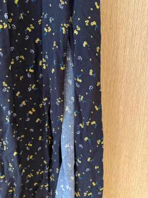 Svart blommig midiklänning H&M - Svart klänning från H&M i storlek XS med små blommor i gult, blått och vitt. Klänningen har långärmade puffärmar, fyrkantig ringning, smockad överdel och slits på sidan. Tillverkad i mjuk viskos som faller snyggt. Blommig långärmad klänning med slits och liten puffärm.