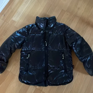 Svart pufferjacka från H&M - Svart, glansig pufferjacka från H&M med Thermolite EcoMade-foder. Jackan har hög krage, två sidofickor med dragkedja och en klassisk quiltad look. Perfekt för kalla dagar och har en modern, loose passform som är både bekväm och snygg.