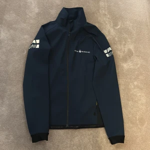 Mörkblå vindjacka från Sail Racing - Snygg mörkblå vindjacka från Sail Racing med vita logotryck på bröstet, ryggen och båda ärmarna. Jackan har hög krage, hel dragkedja och långärmad design. Materialet är slitstark polyester med meshfoder på insidan för extra komfort. Perfekt för dig som gillar sportig stil.