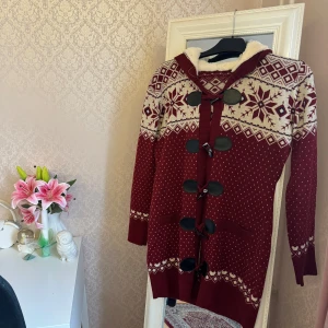Vinröd stickad kofta med huva - Mysig vinröd stickad kofta med huva och vita mönster i nordisk stil. Koftan har stora svarta knappar med snörning framtill, två fickor och är långärmad. Perfekt för kalla dagar och har en riktigt vinter- och julkänsla. Köpt utomlands men bedömer att storleken är i S eller EU 36 då 🤍