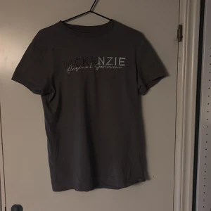 Grå McKenzie t-shirt med broderi - Snygg grå t-shirt från McKenzie med broderad logga och text på bröstet. Klassisk rund hals och korta ärmar. Materialet är mjuk bomull som känns skönt mot huden. Perfekt för dig som gillar stilren streetwear med sportig vibe. Men det finns en liten fläck på tröjan.