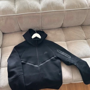Svart Nike tech - Svart vindjacka från Nike med huva och hel dragkedja framtill. Jackan har en diskret logga på bröstet, en snygg diagonal detalj över bröstet och en ficka med dragkedja på ärmen. Materialet är mjukt och känns sportigt, perfekt för chill eller träning. Den är helt ny vill inte har den längre