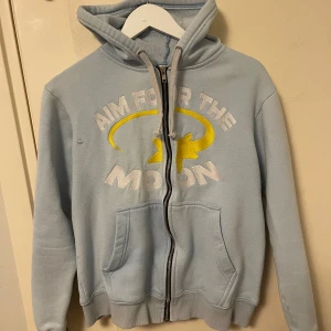 Ljusblå hoodie Aim For The Moon - Ljusblå hoodie från Aim For The Moon med broderad text och gul stjärna på bröstet.OBS DEN PASSAR BÄST FÖR STORLEK S!!!. Tröjan har huva, snörning och dragkedja framtill samt två fickor. Mjuk insida i bomullsmix och ribbade muddar vid ärmslut och nederkant. Perfekt för chill dagar.
