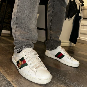 Gucci ace skor  - Snygga vita sneakers från Gucci med klassiska rödgröna ränder på sidorna och broderad guldfärgad bi. Skorna har snörning, platt sula och detaljer i rött och grönt ormskinn på hälen. Tillverkade i skinn och passar perfekt till en trendig style 