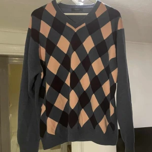 Rutig blå och rosa tröja från Wolsey - Snygg stickad tröja från Wolsey i blått med rutigt mönster i rosa och mörkblått framtill. V-ringad hals och långa ärmar. Perfekt för dig som gillar preppy stil och vill sticka ut lite extra med färg och mönster.