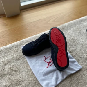 Svarta sneakers från Christian Louboutin - Snygga svarta sneakers från Christian Louboutin med klassisk röd sula och diskret logga. Skorna har rund tå, snörning och är tillverkade i textil. Perfekta för dig som vill ha en lyxig och stilren look.