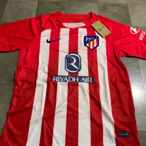 Atletico Madrid randig fotbollströja Nike - Säljer en officiell Atletico Madrid fotbollströja från Nike i storlek S. Tröjan är röd och vit med vertikala ränder, klubbmärke på bröstet och sponsorer som Riyadh Air och Hyundai. Tillverkad i lätt polyester med korta ärmar och rund hals.