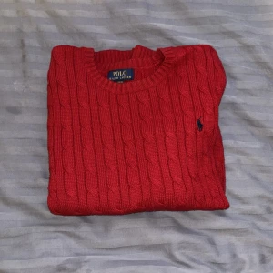 Röd stickad tröja Polo Ralph Lauren - Snygg röd stickad tröja från Polo Ralph Lauren med klassiskt kabelstickat mönster och rund hals. Tröjan har en liten broderad logga på bröstet och ribbade muddar vid hals, ärmslut och nederkant. Perfekt för dig som gillar stilrena och tidlösa plagg.