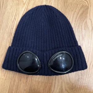 Mörkblå C.P. Company mössa   - Ribbstickad mössa i mörkblått från C.P. Company med ikoniska svarta goggles framtill på uppviket. 100% ull. Snygg och unik design som sticker ut. Köpta här på Plick men används ej längre 