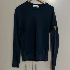 Marinblå sweatshirt från Stone Island - Snygg marinblå sweatshirt från Stone Island med klassisk rund hals och lång ärm. Ikonisk logotyp-patch på vänster ärm med gul och grön brodering. Tröjan har ribbade muddar och är tillverkad i mjuk bomull för skön komfort.