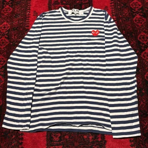 Randig långärmad tröja Comme des Garçons - Säljer en ikonisk långärmad tröja från Comme des Garçons PLAY i marinblått och vitt med randigt mönster. Tröjan har ett rött hjärta med ögon broderat på bröstet och rund hals. Perfekt för dig som gillar streetstyle och vill sticka ut.