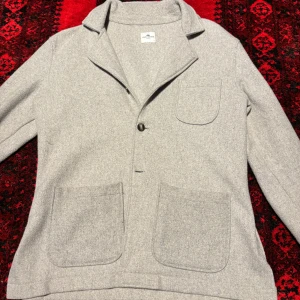 Grå cardigan jacka från John Henric - Snygg ljusgrå cardigan jacka från John Henric med klassisk krage och tre stora fickor framtill. Jackan har en enkel knäppning med knapp och är tillverkad i mjuk ull som ger en chill och clean look. Perfekt för lager-på-lager och stilrena outfits.