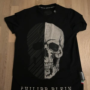 Svart Philipp Plein t-shirt med dödskalle - Svart t-shirt från Philipp Plein med stor dödskalle framtill, där ena halvan är randig och andra halvan är dekorerad med små paljetter för extra bling. Klassisk passform och rund hals. Märkesdetaljer syns i nacken och på sidan. Perfekt för dig som gillar edgy stil. Skriv gärna innan köp!!!🔥🔥🔥