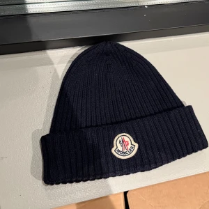 Mörkblå ribbad mössa från Moncler - Snygg mörkblå mössa från Moncler med ribbad stickning och uppvikt kant. Märkets ikoniska logotyp är broderad framtill. Tillverkad i mjuk ull som håller dig varm och ger en clean look. Perfekt för dig som vill ha både stil och komfort.