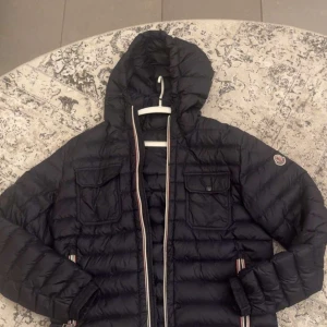Svart Moncler pufferjacka med huva - Svart pufferjacka från Moncler med huva och klassisk logga på ärmen. Jackan har två bröstfickor med lock, dragkedja framtill med röd-vita detaljer och quiltad design. Insidan har märkning 'Longue Saison'. Perfekt för dig som vill ha en snygg och varm jacka.