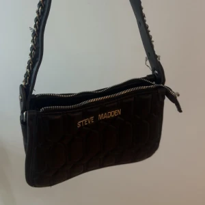 Svart quiltad axelväska Steve Madden - Snygg svart quiltad axelväska från Steve Madden med kedjedetaljer på axelremmen och guldfärgad logga framtill. Väskan har dragkedja upptill och en cool struktur i materialet som ger extra edge till din outfit.