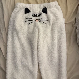Vita fluffiga mjukisbyxor med kattmotiv - Supergosiga vita mjukisbyxor med broderat kattansikte och öron på baksidan, samt texten 'SLEEPY'. Byxorna har en ljusrosa satinrosett framtill och är tillverkade i mjukt fleece-material. Perfekta för chill hemma och mysiga dagar.