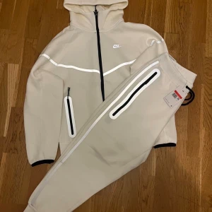 Vit Nike reflective tech tracksuit  - Snygga vit mjukisbyxor från Nike med svarta och vita reflexdetaljer längs benen. Byxorna har dragkedjor vid fickorna och en modern, sportig look. Perfekt för dig som gillar streetwear och vill sticka ut med coola detaljer.