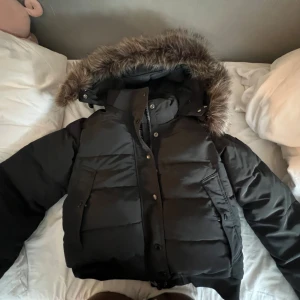 Svart pufferjacka Superdry Everest - Svart pufferjacka från Superdry Everest med stor huva och fluffig fuskpäls. Jackan har flera fickor framtill, Superdry-logga på ärmen och dragkedja med knappar. Perfekt för kalla dagar med sin tjocka vaddering och snygga look.Pris kan diskuteras!! 