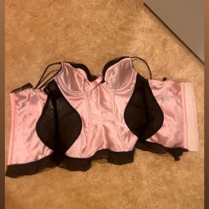 Rosa och svart korsett från Victoria's Secret - Supersnygg rosa korsett från Victoria's Secret med svarta meshdetaljer och liten rosett framtill. Satinliknande material och croppad modell med tunna axelband. Perfekt för dig som vill sticka ut med en feminin och edgy vibe.