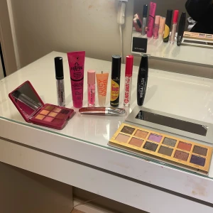 Sminkpaket med paletter och gloss - Säljer ett sminkpaket med W7 Moonglade ögonskuggspalett med 12 färger i nude, lila och guld, en mindre röd ögonskuggspalett med rosa och bruna toner, Technic Mega Lash mascara, Essence Get Big! Lashes mascara, Hot Shot eyeliner, Dr.PAWPAW balm, två läppglans (Glitter Gloss och Juicy Bomb) och Pink Sparks gloss. Dem flesta sakerna är lite använda men rätt så nya☺️