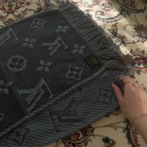 Grå Louis Vuitton halsduk med monogram - Snygg grå halsduk från Louis Vuitton med ikoniskt monogrammönster och fransar i kanterna. Gjord i 94% ull och 6% silke, vilket gör den både mjuk och lyxig. Perfekt accessoar för att höja din stil under kyliga dagar.