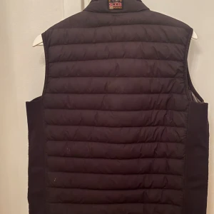 Svart dunväst från Parajumpers - Snygg svart dunväst från Parajumpers med quiltad design och dragkedja framtill. Insidan är ljusgrå och västen har två praktiska fickor med dragkedja. Perfekt att slänga över en hoodie för extra värme. Klassisk look med hög krage.