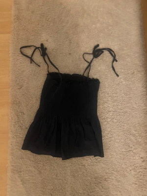 Svart smockad linnetopp med knytband - Svart linnetopp med smockad överdel och volangig nederdel. Toppen har tunna axelband som knyts upptill för en söt och justerbar passform. Perfekt till varma dagar och enkel att styla med jeans eller kjol.