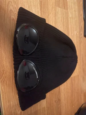 Svart Goggle-mössa från C.P. Company - Säljer nu min CP Company mössa som bara blivit använd ett fåtal gånger och därför är i riktigt fint skick. Den är väl omhändertagen, helt utan skador eller slitningar, och ser i princip ny ut. Perfekt för dig som vill ha en stilren och kvalitetsmössa utan att betala fullpris i butik. Passar till alla huvuden.   Mössan är äkta CP Company och har den klassiska, clean designen som märket är känt för. Passar till både vardag och mer uppklädda outfits. Kommer från ett rök- och djurfritt hem. 