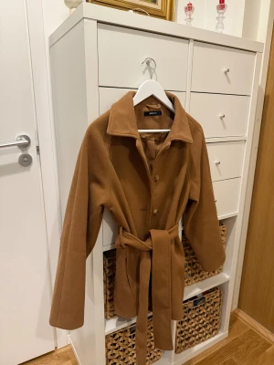 Camelbrun kappa från Gina Tricot - Stilren camelbrun kappa från Gina Tricot med klassisk krage, knappar framtill och matchande bälte i midjan. Jackan har långa ärmar och en rak, något loose passform. Perfekt för dig som vill ha en tidlös look med snygg siluett.