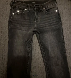 Svarta jeans från True Religion - Säljer ett par svarta jeans från True Religion med klassiska sömmar och silverfärgade knappar på bakfickorna. Jeansen har utsvängda ben 