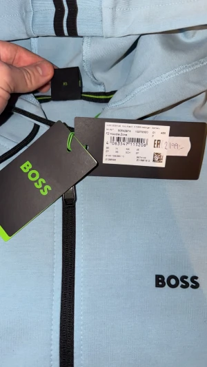 Ljusblå hoodie från BOSS med dragkedja - Snygg ljusblå hoodie från BOSS med svart dragkedja och logga på bröstet. Tröjan har huva med snörning och svarta detaljer på huvan. Perfekt för dig som gillar stilrena och sportiga plagg. Materialet är mjukt och känns skönt mot huden. Hoodien är helt ny, från urban modern i Halmstad, använd 1 gång och passade inte min stil, pris kan diskuteras!😃