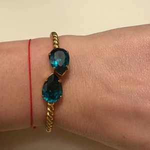 Guldfärgat armband Caroline Svedbom - Snyggt armband från Caroline Svedbom i guldfärgad metall med tvinnad design och två stora, droppformade stenar. Statement-piece som verkligen drar blickarna till sig och ger en lyxig känsla till din outfit.