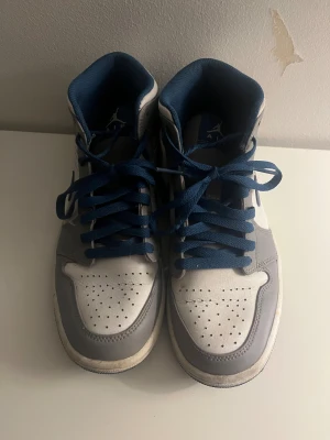 Nike Air Jordan 1 Mid grå/blå - Nike Air Jordan 1 Mid i grått och vitt med blå detaljer och snörning. Klassisk siluett med blå swoosh och blå innersula. Ovandel i skinn och syntet, perforerad tå och vit mellansula. Perfekta för dig som gillar streetwear och sneakers med retrovibe.
