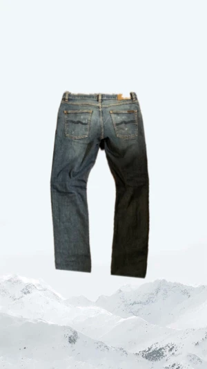  Nudie Jeans  - Säljer ett par blå slim fit jeans från Nudie Jeans med slitna detaljer och klassisk femficksdesign. Jeansen har en mörkblå våg på bakfickorna och läderpatch med logga i midjan. Materialet är jeans med snygg tvätt och slitningar för en cool look.