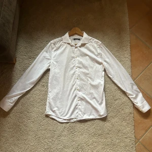 Vit skjorta från J.Lindeberg - Klassisk vit skjorta från J.Lindeberg med snygg krage och knappar framtill. Skjortan har lång ärm och är tillverkad i mjuk bomull som känns skön mot huden. Perfekt för dig som gillar stilrena och enkla plagg med bra passform.