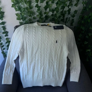 Vit kabelstickad tröja Polo Ralph Lauren - Vit kabelstickad tröja från Polo Ralph Lauren med klassisk rund hals och broderad logga på bröstet. Tröjan har ribbade muddar vid ärmslut och nederkant, och är tillverkad i mjuk bomull. Perfekt för dig som gillar stilrena och tidlösa plagg.