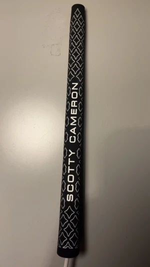 Scotty Cameron Putter Grip - Snygg och bekväm Scotty Cameron puttergrepp i svart med vit mönsterdesign. Greppet är i mycket gott skick utan synligt slitage eller defekter. Perfekt för dig som vill uppgradera din golfputter med ett premiumgrepp från en ledande tillverkare.