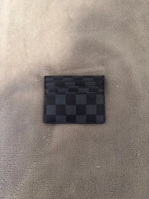 Svart rutig korthållare - Slimmad och stilren kortplånbok från ni vet vart i svart och grått med klassiskt rutigt Damier-mönster. Plånboken är tillverkad i skinn och har flera fack för kort. Perfekt för dig som vill ha en lyxig och praktisk accessoar i fickan.