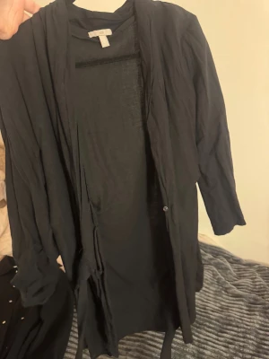 Svart cardigan jacka från H&M - Svart lång cardigan jacka från H&M med bälte i midjan och långa ärmar. Jackan är tunn och har en avslappnad passform, perfekt att slänga över en t-shirt eller hoodie. Materialet känns mjukt och lätt, vilket gör den enkel att bära hela dagen.