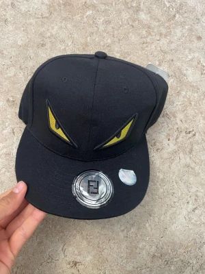 Svart Fendi keps med gula monsterögon - Säljer en svart keps från Fendi med broderade gula monsterögon framtill och Fendi-logga bak. Kepsen har klassisk form, böjd skärm och justerbar snapback. Tillverkad i bomull och har coola detaljer som gör den till ett statement-piece.